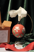 Load image into Gallery viewer, Round cinnamon-red Christmas ornament hand-painted with cream winter jasmine flowers, hanging from a brass stand beside its kraft gift box