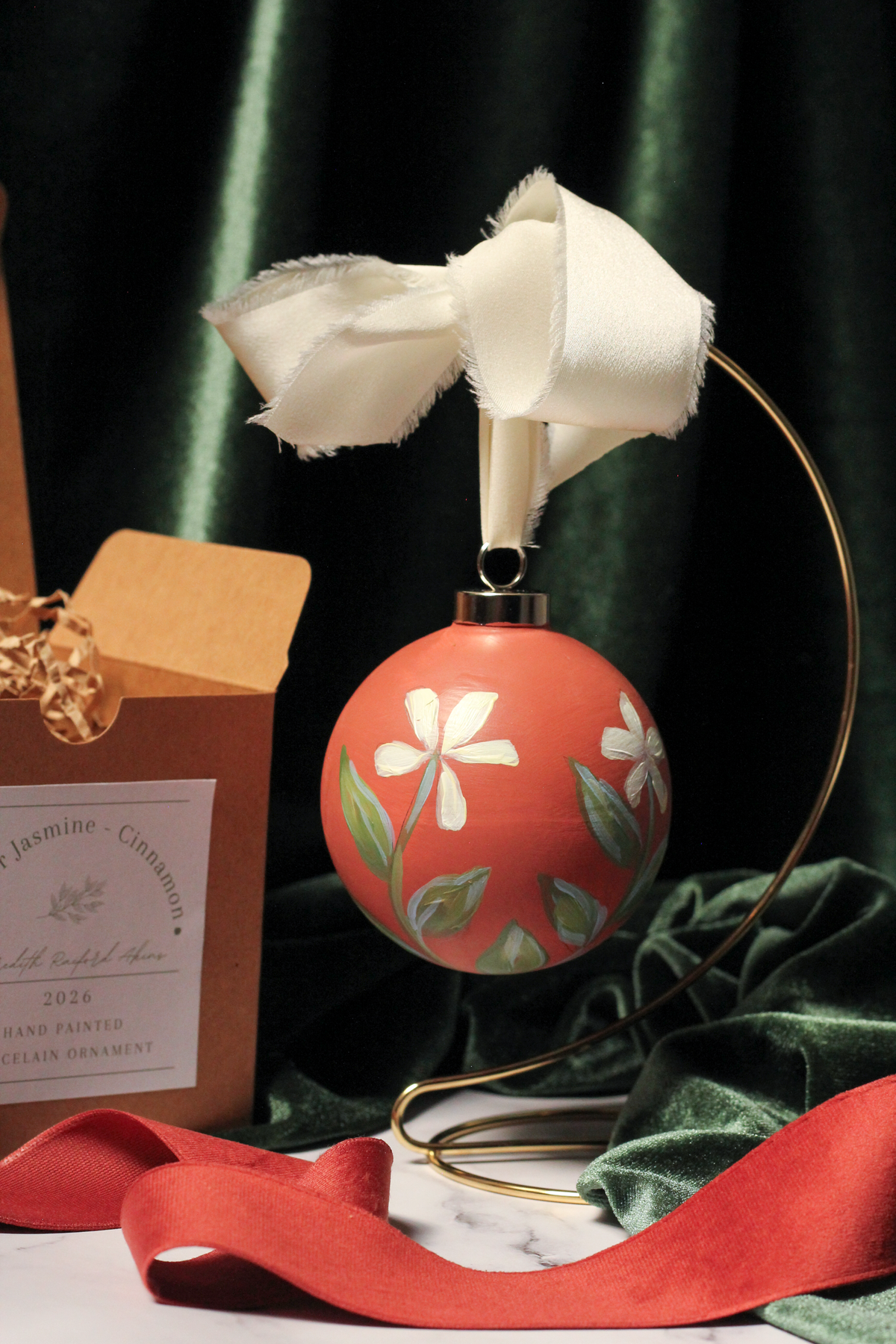 Round cinnamon-red Christmas ornament hand-painted with cream winter jasmine flowers, hanging from a brass stand beside its kraft gift box