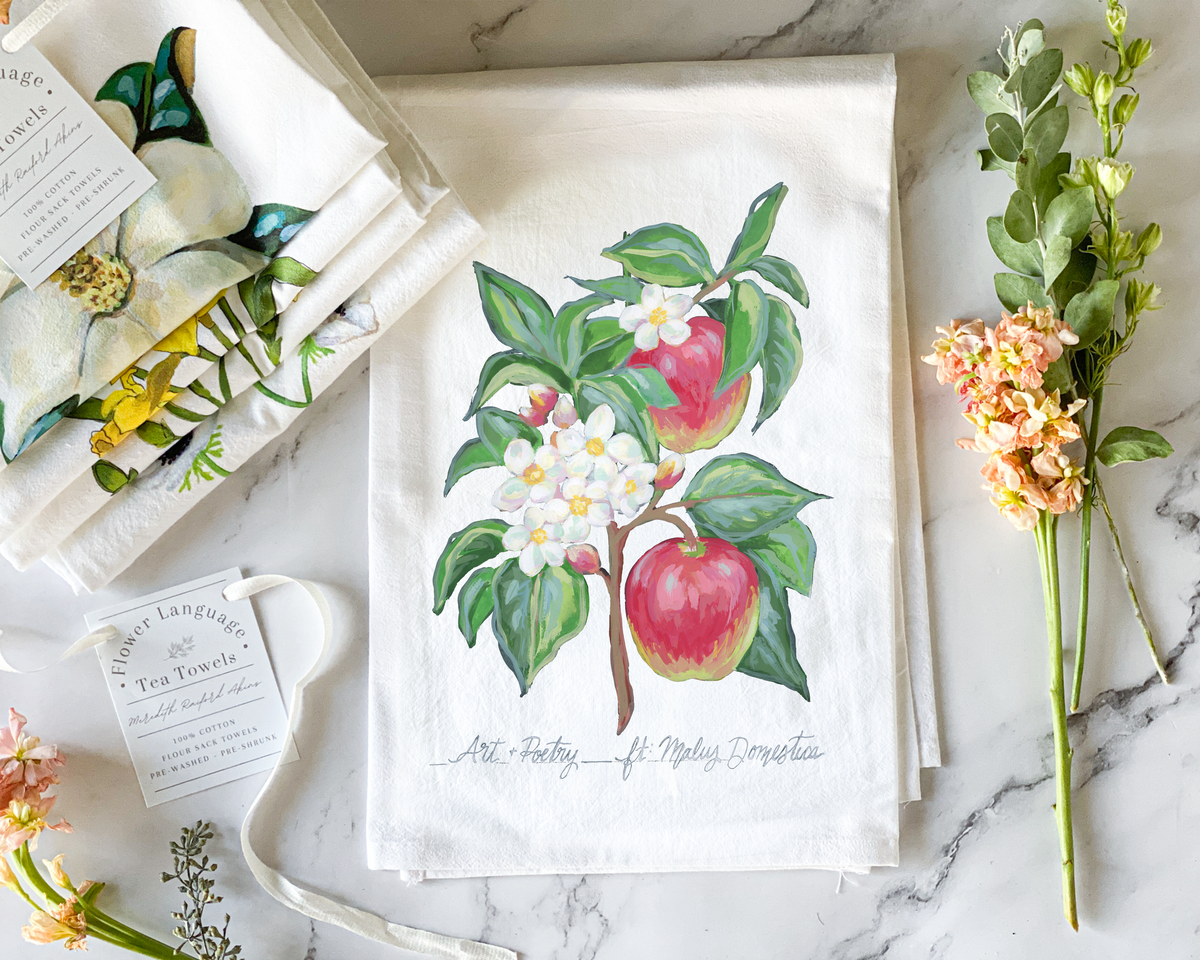 White cotton flour sack tea towel with a hand-painted watercolor apple tree branch, red apples and white blossoms, lettered with its Victorian flower meaning Art and Poetry, styled on marble beside folded botanical tea towels and fresh flowers.