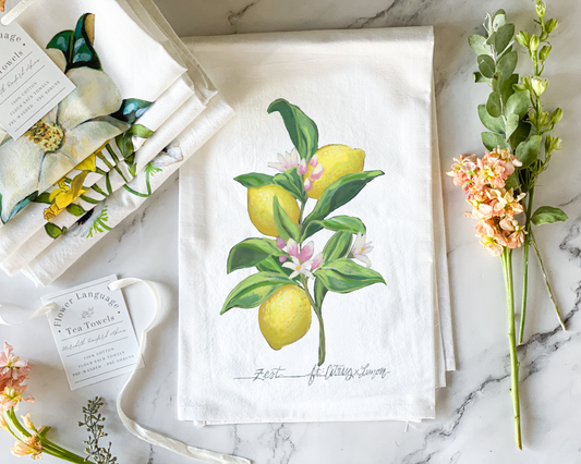 White cotton flour sack tea towel with a hand-painted watercolor lemon tree branch, yellow lemons and pink-tinged blossoms, lettered with its Victorian flower meaning Zest, styled on marble beside folded botanical tea towels and fresh flowers.