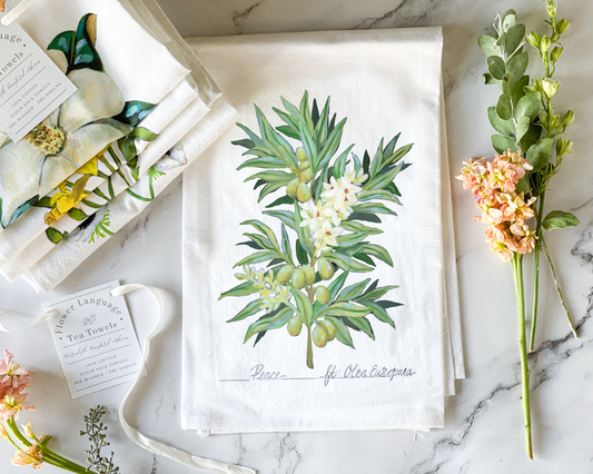 White cotton flour sack tea towel with a hand-painted watercolor olive branch, green olives and cream blossoms, lettered with its Victorian flower meaning Peace, styled on marble beside folded botanical tea towels and fresh flowers.