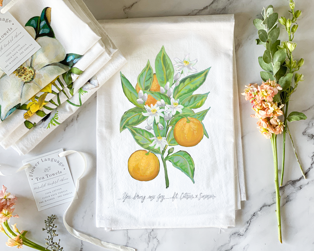 White cotton flour sack tea towel with a hand-painted watercolor orange tree branch, ripe oranges and white orange blossoms, lettered with its Victorian flower meaning You Bring Me Joy, styled on marble beside folded botanical tea towels and fresh flowers.