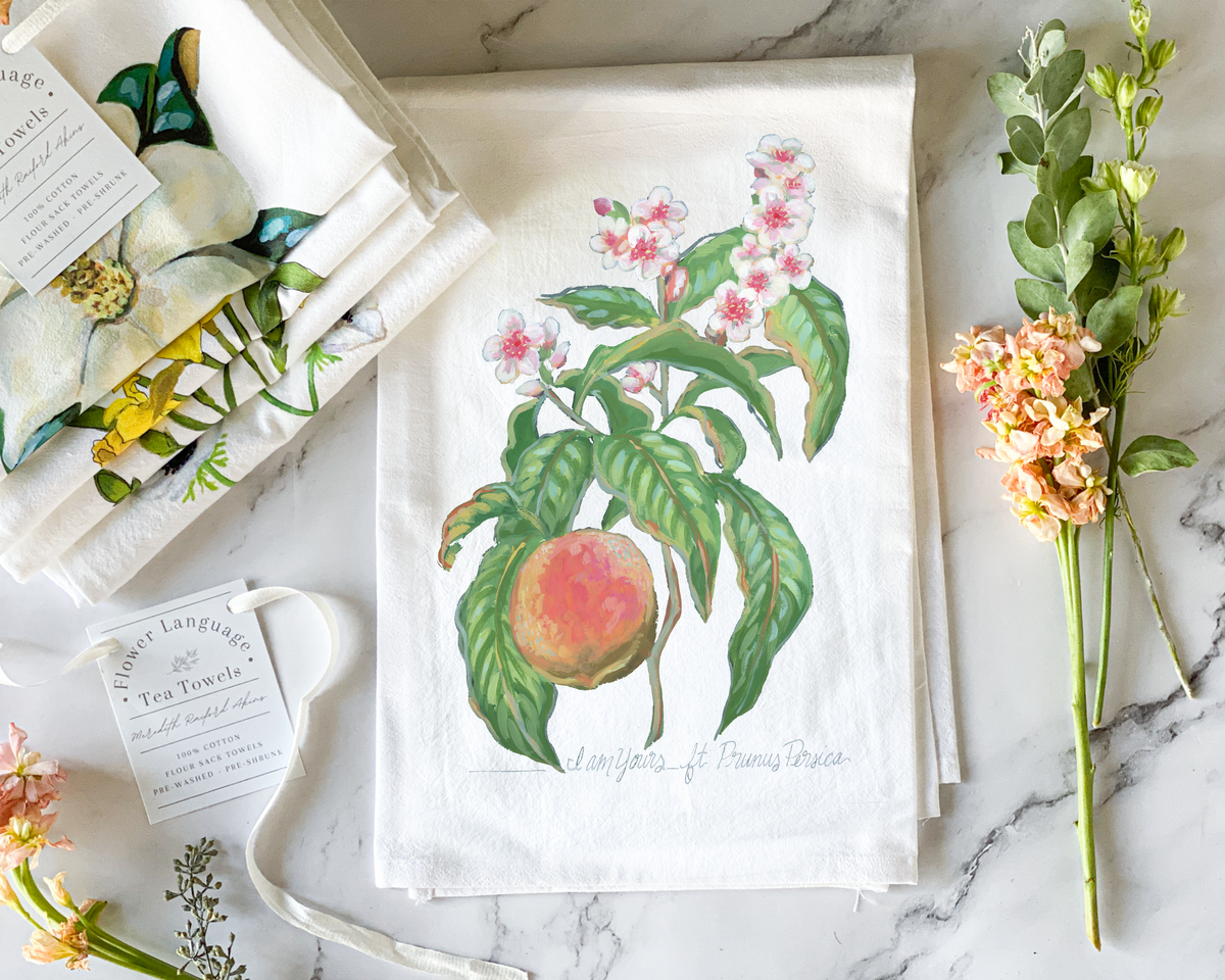 White cotton flour sack tea towel with a hand-painted watercolor peach tree branch, a ripe peach and pink blossoms, lettered with its Victorian flower meaning I Am Yours, styled on marble beside folded botanical tea towels and fresh flowers.