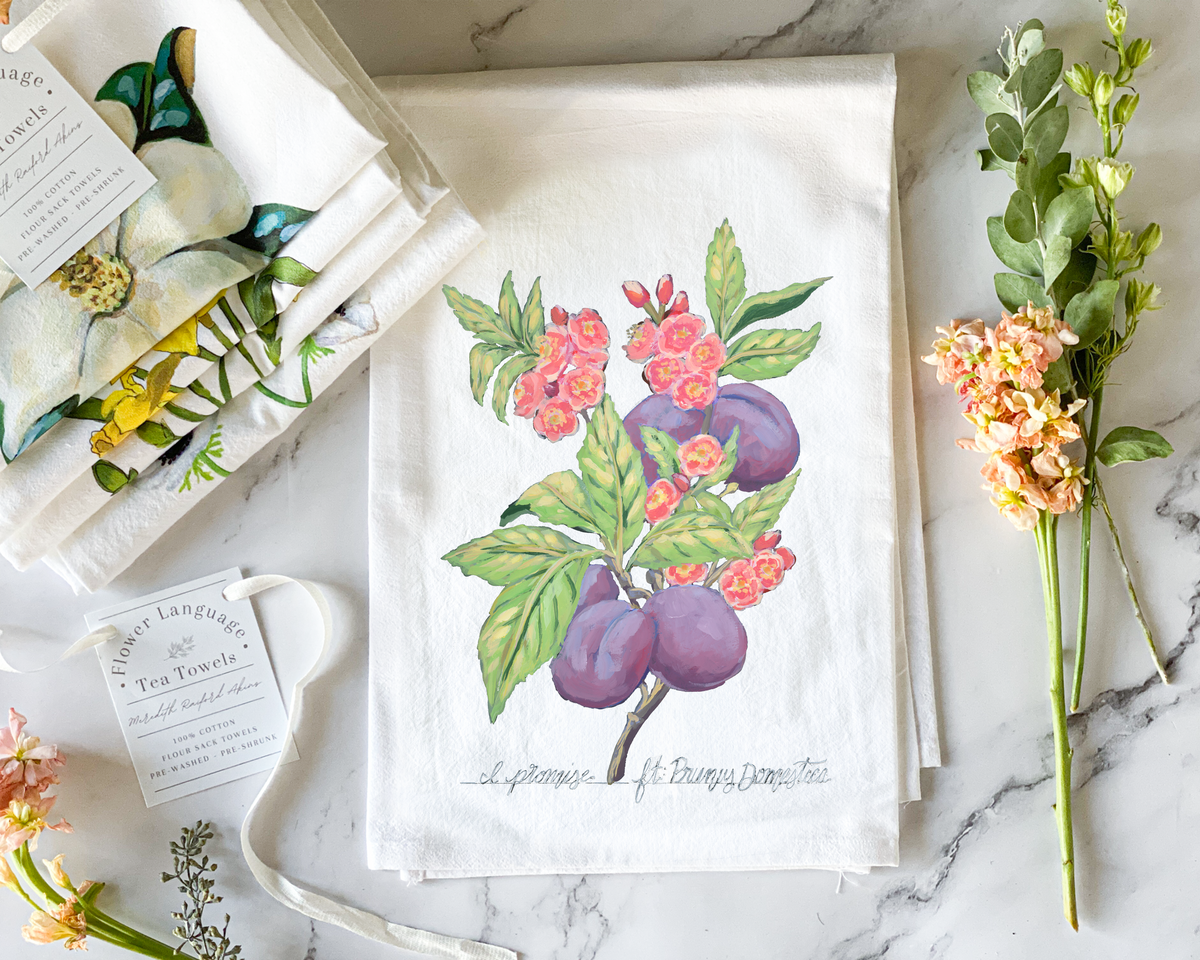 White cotton flour sack tea towel with a hand-painted watercolor plum tree branch, purple plums and pink blossoms, lettered with its Victorian flower meaning I Promise, styled on marble beside folded botanical tea towels and fresh flowers.